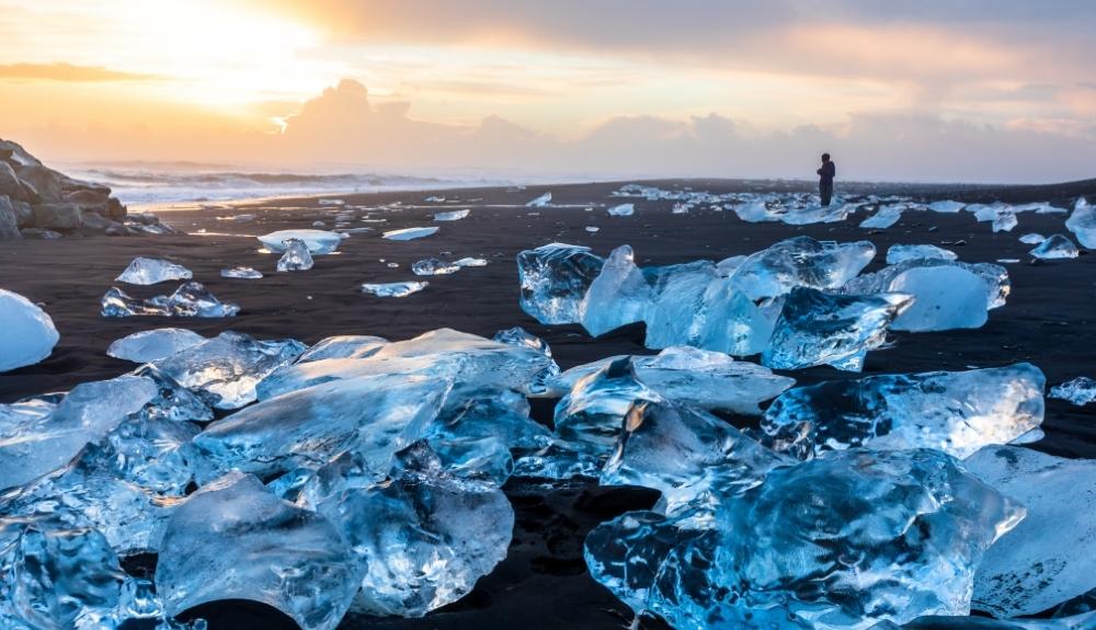 Small glacier chunks on a black sand beach all throughout the photo. A person is standing among them, the sun is setting. The ocean is slightly in the distance of the image. Diamond Beach in Iceland