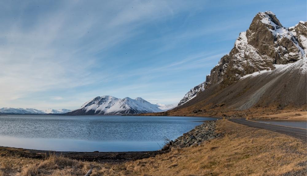 A body of water in the foreground on the left with a road in the foreground on the right, snowcapped mountains in the background. A picture of Iceland.