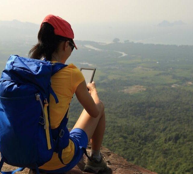 Hiker wearing a blue backpack is reading on the edge of a cliff in the foreground, a large dropoff of greenery in the background. Emphasis on the blog post books for hikers.