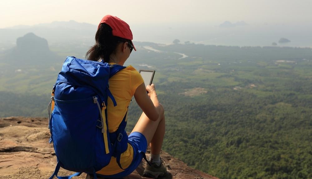 Hiker wearing a blue backpack is reading on the edge of a cliff in the foreground, a large dropoff of greenery in the background. Emphasis on the blog post books for hikers.