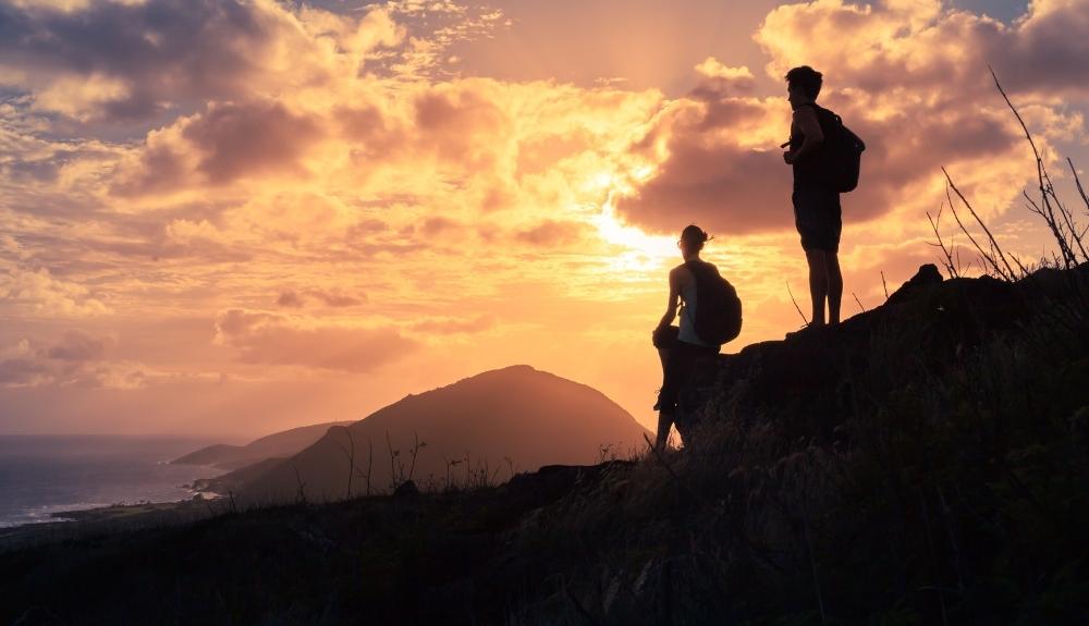 Silhouette of two hikers on a cliff's edge in the foreground, a setting sun in the background. 