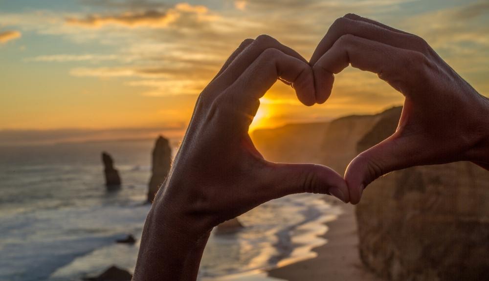 A pair of hands creating a heart in the foreground, a beach and sunset in the background. Emphasis on self care for travel essentials for women.