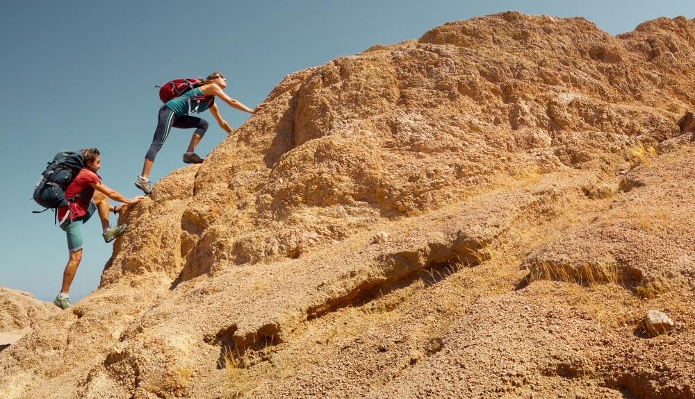 Two hikers on the left are struggling to climb up a rock formation that is to the right. Emphasis on books for hikers